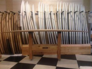Varias tablas de surf están apiladas verticalmente contra una pared de madera en Core Surfing Shop, con una tabla larga exhibida horizontalmente sobre un soporte de madera en primer plano. El suelo tiene un patrón de cuadros en blanco y negro.