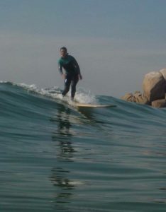 Un surfista cabalga una ola sobre una tabla de surf Firewire TJ Everyday amarilla y aparece en equilibrio con las rodillas dobladas en un mar en calma, mientras que se pueden ver grandes rocas al fondo.