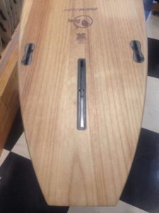 Una tabla de surf Firewire TJ Everyday de madera con tres ranuras para quillas descansa sobre un suelo cuadriculado. La insignia en el tablero dice "FIREWIRE", "TIMBERTEK" y "X Surfing Games".