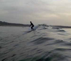 Un surfista con traje de neopreno monta una ola creciente, manteniendo el equilibrio sobre la tabla de surf, en un paisaje marino nublado cerca de una costa lejana.