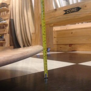 Una cinta métrica se encuentra en posición vertical sobre un piso a cuadros cerca de muebles de madera con la etiqueta "CORE". La cinta se extiende verticalmente junto a una tabla de surf de madera hasta los 4 centímetros