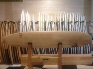 Una tabla de surf de madera descansa sobre un soporte de madera en nuestra tienda de surf, rodeada de varias tablas de surf blancas y de madera alineadas contra una pared de madera. El texto en el stand dice "CORE".