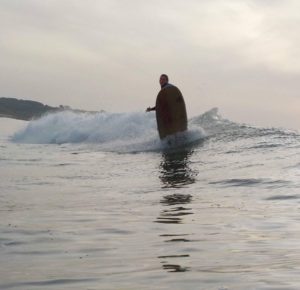 Un surfista cabalga una pequeña ola con un ligero giro mientras está parado sobre una tabla de surf, rodeado por el mar abierto y bajo un cielo nublado. Al fondo se ven colinas lejanas.