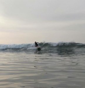 Un surfista cabalga una ola de tamaño mediano, haciendo equilibrio sobre la tabla de surf, con un mar en calma y un cielo nublado de fondo.