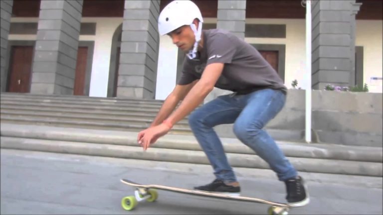 Luis Rua de Miller Division con un casco blanco agachado sobre un skate, paseando por una acera gris junto a un edificio con grandes columnas y escalones de piedra.