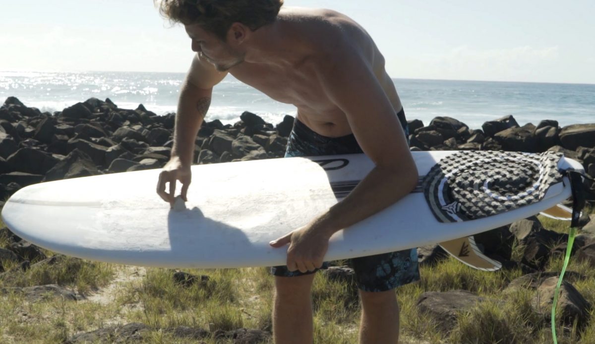 Tomo sin camisa encerando una tabla de surf Slater Designs Omni en una costa rocosa y cubierta de hierba, con el océano al fondo bajo un cielo despejado en Lennox Head