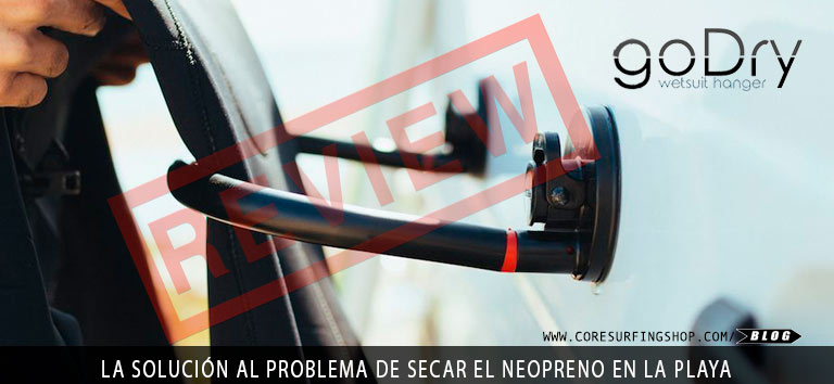 Se está colocando una percha negra para trajes de neopreno en la ventana de un vehículo. El texto lee: "percha para traje de neopreno goDry". El texto rojo superpuesto dice: "REVIEW". Debajo dice: "LA SOLUCIÓN AL PROBLEMA DE SECAR EL NEOPRENO EN LA PLAYA".