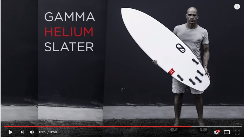 Kelly Slater sostiene una tabla de surf blanca frente a una pared oscura. El texto en la pared dice "GAMMA HELIUM SLATER"