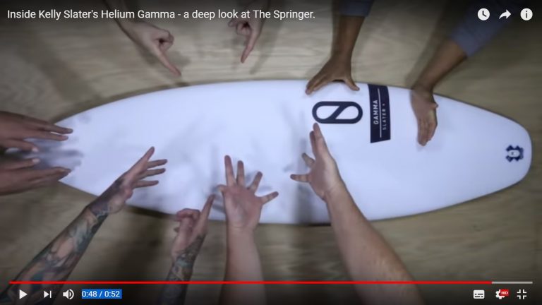 Una tabla de surf springer slater gamma helium es examinada por varias manos que apuntan a varias partes, colocadas sobre un piso de madera. El texto "Dentro de Helium Gamma de Kelly Slater: una mirada profunda a The Springer" está en la parte superior.