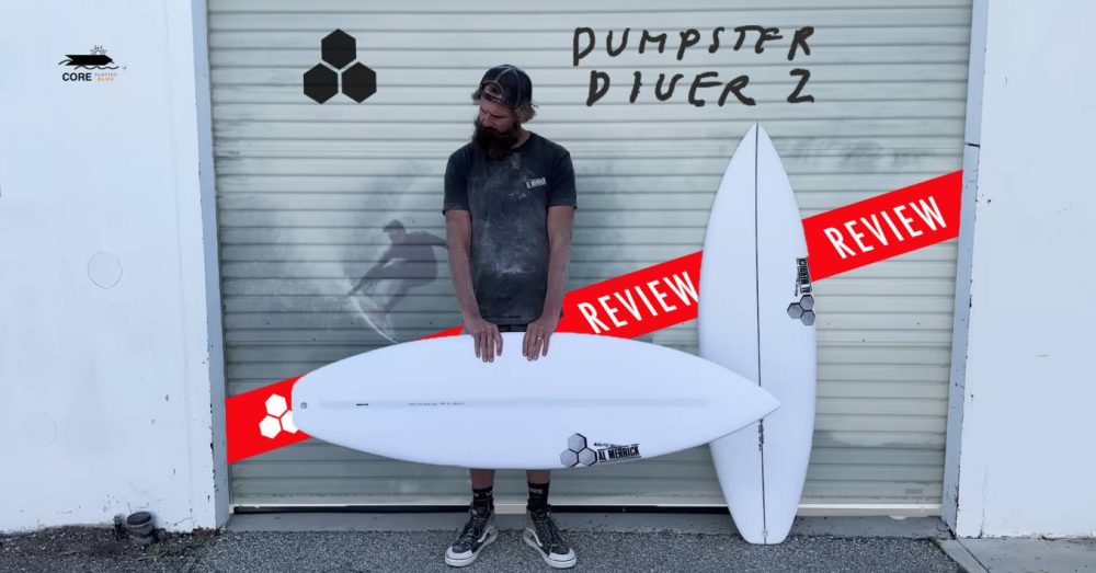 Un hombre está de pie y sostiene una tabla de surf CI Dumpster Diver blanca afuera junto a otra tabla de surf contra una puerta enrollable de color beige con "DUMPSTER DIVER 2" escrito en negro y el texto "REVIEW" en una pancarta roja.